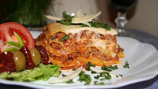Kousek pečené lasagne s rajčaty a olivami na talíři.
