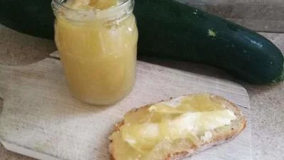 Sklenice cuketové marmelády s kouskem chleba vedle čerstvého okurky.