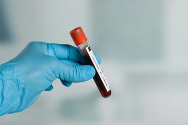 Ruka v modrých rukavicích drží zkumavku s krví pro laboratorní vyšetření.