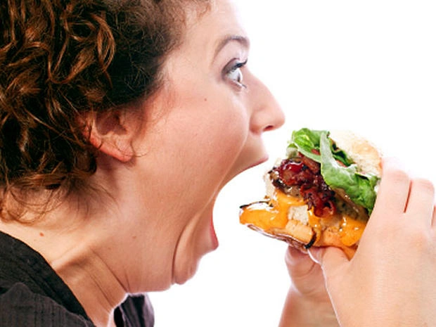 Žena se snaží sníst velký hamburger s listovou zeleninou.