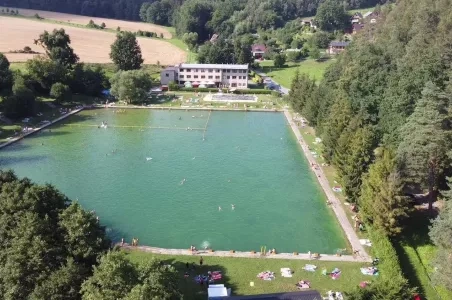 Venkovní bazén v přírodě s lidmi plavajícími a odpočívajícími.