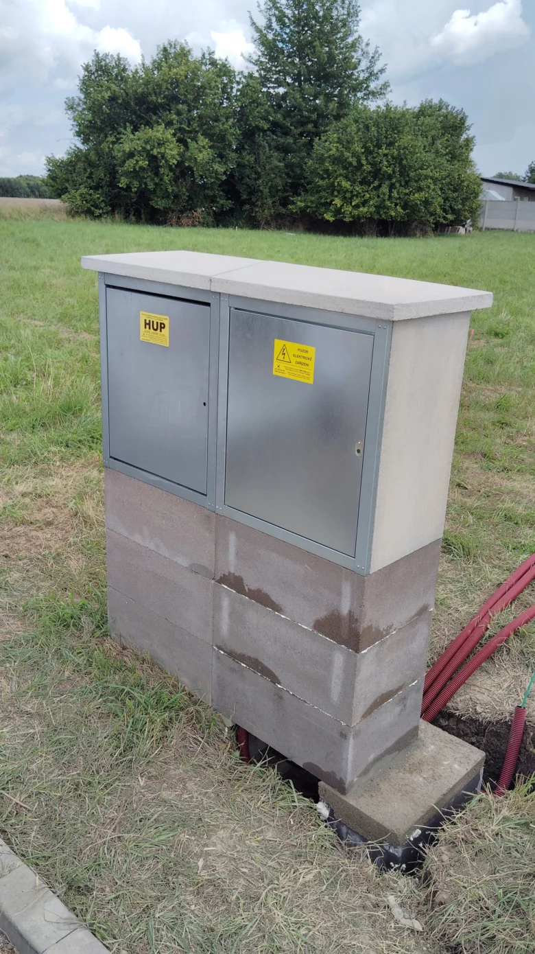 Šedý elektrický rozvaděč na betonu v poli s červenými kabely.
